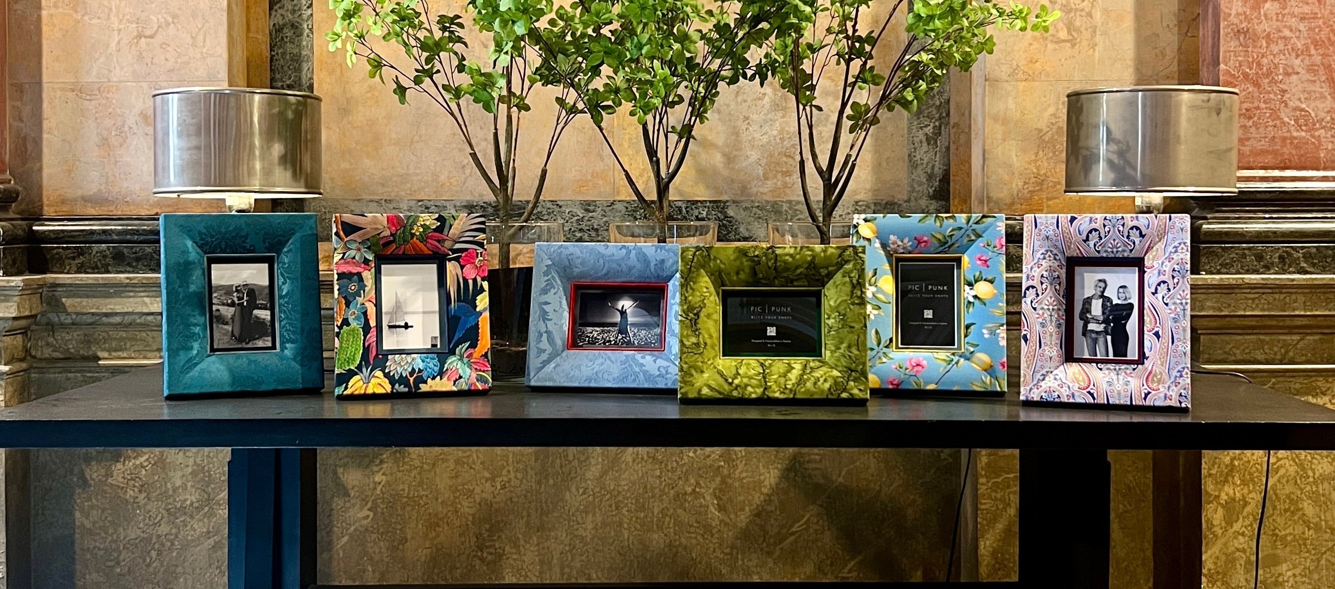 Sechs stoffbezogene Designer-Bilderrahmen von PIC | PUNK® auf einem schwarzen Tisch in einem historischen Foyer, flankiert von zwei Lampen und frischem Grün – eine stilvolle Komposition aus Handwerk, Textil und Architektur.