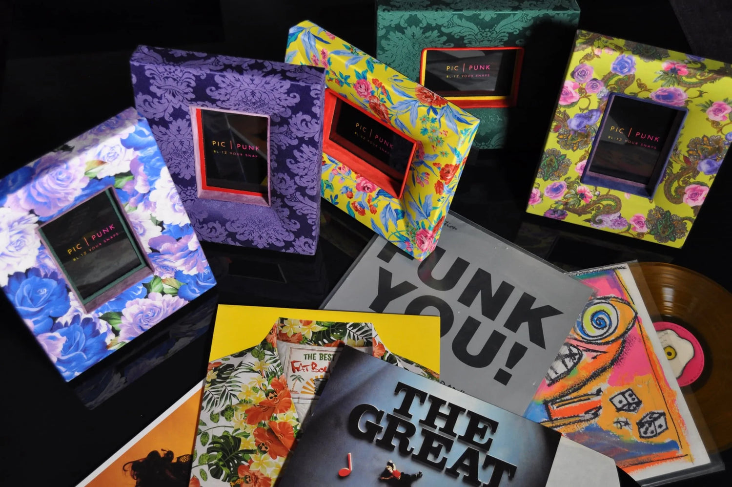 Stoffbezogene Bilderrahmen von PIC | PUNK auf einem schwarzen Glastisch, arrangiert mit bunten Schallplattenhüllen und Vinyls. Die einzigartigen Rahmen aus Fichtenholz sind mit floralen Stoffen bezogen und treffen auf musikalische Inspiration.