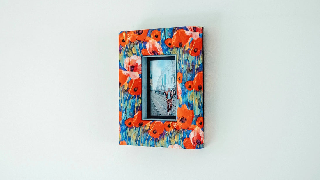 Stoffbezogener Bilderrahmen mit lebhaftem Mohnblumenmuster in Rot und Blau, aufgehängt an einer weißen Wand. Im Rahmen ein Foto mit Straßenmotiv. Der Rahmen wirkt modern und kunstvoll.