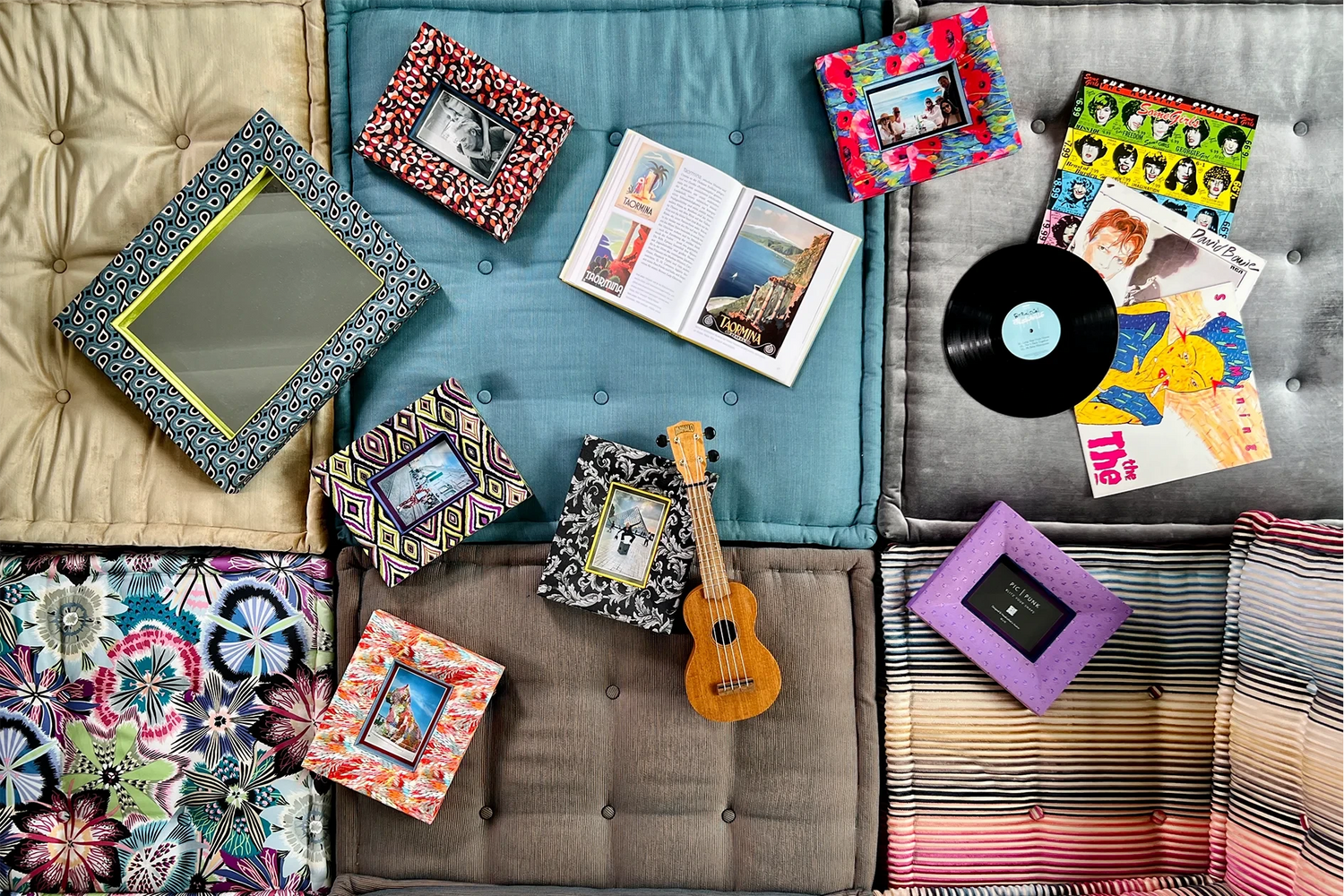 Bunte, stoffbezogene Bilderrahmen und ein Spiegel liegen locker arrangiert auf einem modernen Sofa mit verschiedenfarbigen Kissen. Dazwischen eine kleine Ukulele, eine geöffnete Zeitschrift und Schallplatten – ein künstlerisches, verspieltes Interior-Setting.