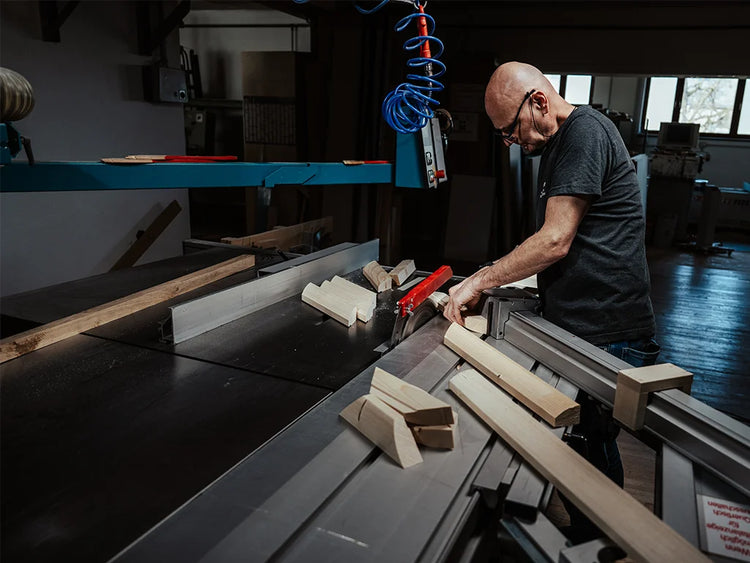 Ein Tischler fertigt in einer österreichischen Werkstatt Rohrahmen aus Fichtenholz – die Grundlage für unsere stoffbezogenen Bilderrahmen. Präzise bearbeitet er die Holzteile mit einer Formatkreissäge.