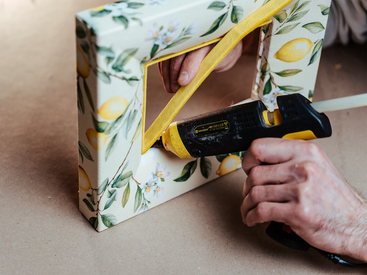 Ein Tapezierer befestigt ein gelbes Samt-Inlay mit einer Heißklebepistole in einem stoffbezogenen Bilderrahmen mit Zitronenmotiv. Die Nahaufnahme zeigt den präzisen Arbeitsschritt beim Veredeln des buttergelben Rahmens.