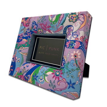 Ein farbenfroher Bilderrahmen im Boho-Stil steht leicht geneigt und hält ein Porträt im Format 10×15 cm. Das lebendige Paisley-Muster in Rosa, Blau und Grün und bringt Bewegung und Wärme in den Raum. Der blaue Innenrand lenkt den Blick auf das Foto und lässt es ruhig wirken, während der gemusterte Rahmen selbst zum dekorativen Element wird. Ideal für offene Regale, Sideboards oder Nachttische in kreativen Wohnkonzepten, die Farbe und Persönlichkeit zeigen wollen.