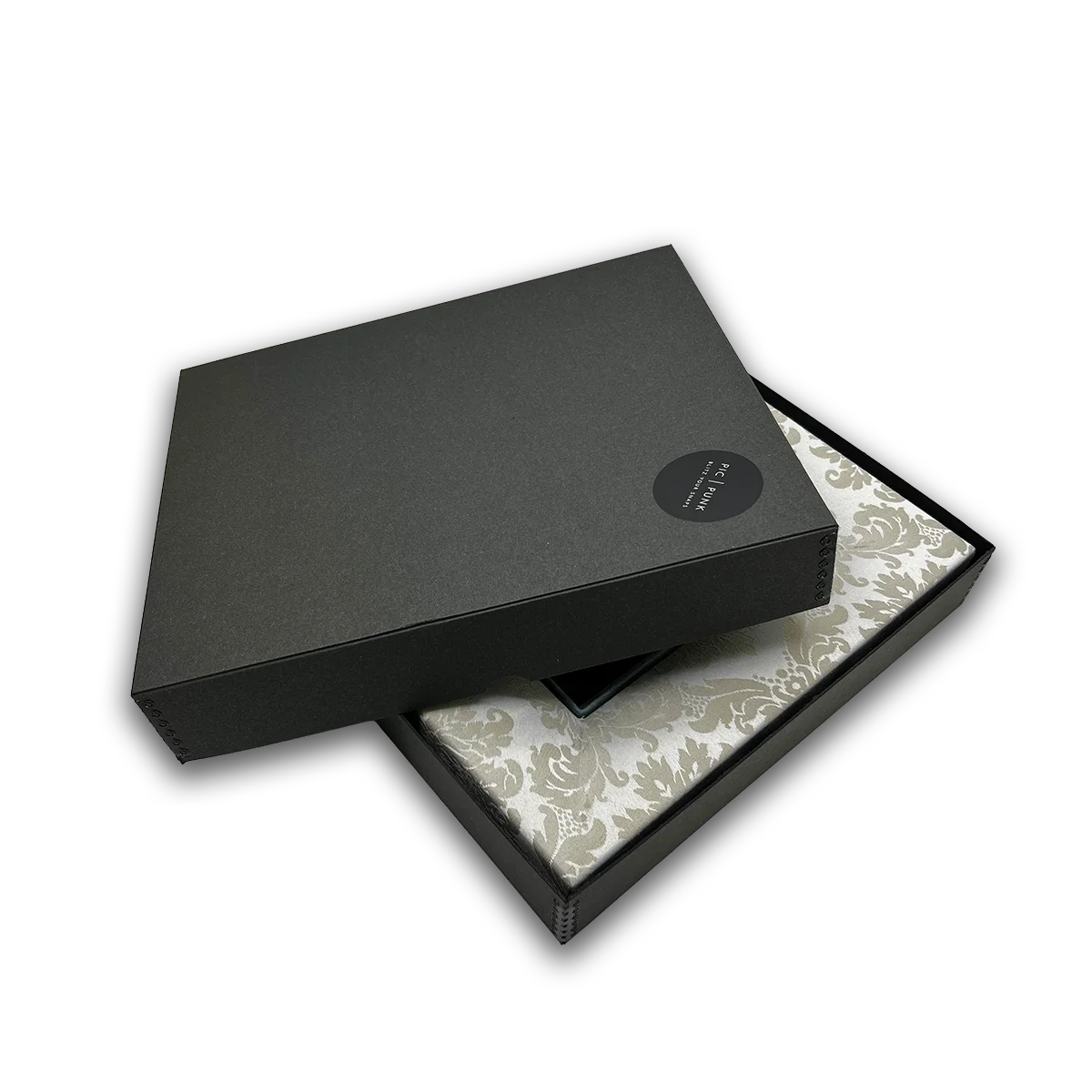 Offene, schwarze Geschenkbox von PIC | PUNK® mit eingelegtem Design-Bilderrahmen „East End Whisper“, hochwertige Verpackung mit passgenauer Form, mattes Kartonmaterial mit dezentem Markenetikett, der elegante Stoffrahmen mit Damastmuster ist teilweise sichtbar, stilvolle Präsentation für handgefertigte Bilderrahmen, ideal als exklusives Geschenk für Interior-Liebhaber:innen, Design-Objekte und besondere Anlässe, gefertigt in Österreich.