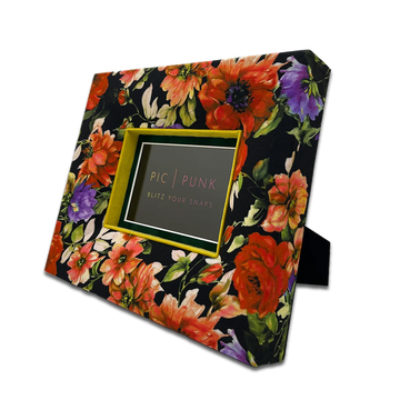 Schräg aufgestellter, handgefertigter Design-Bilderrahmen „Rosy Radiance“ von PIC | PUNK mit üppigem floralen Stoffmuster in Rot-, Orange- und Violetttönen auf schwarzem Grund. Veredelt mit leuchtend grünem Samtpassepartout und tiefem Innenausschnitt. Hochwertiger Stoffrahmen für 10 × 15 cm Fotos, als stilvolles Wohnaccessoire oder Geschenkidee - nachhaltig gefertigt in Österreich.