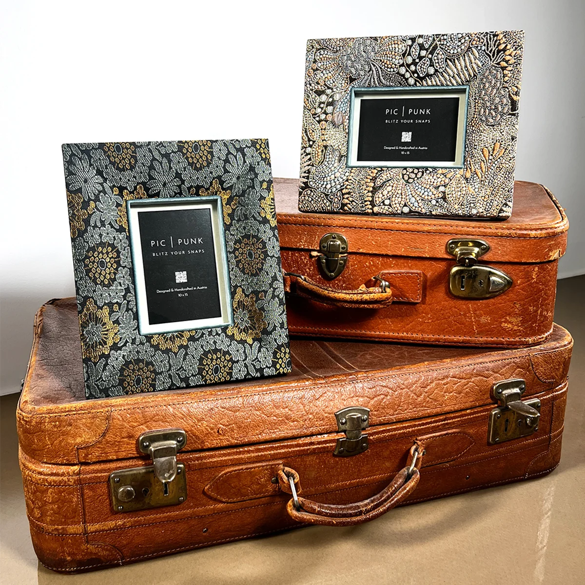 Interior-Fotografie mit zwei edlen Design-Bilderrahmen von PIC | PUNK® auf übereinander gestapelten braunen Lederkoffern: ein rechteckiger Hochformatrahmen und ein Querformatrahmen mit aufwendig gemustertem Stoff in dunklen Grau-, Gold- und Kupfertönen, beide mit markanter Tiefe, farbigem Innenrand und Passepartout, inszeniert als hochwertiges Wohnaccessoire für elegante Raumgestaltung, Boutique-Interiors und ausdrucksstarke Wanddeko, handgefertigt in Österreich.