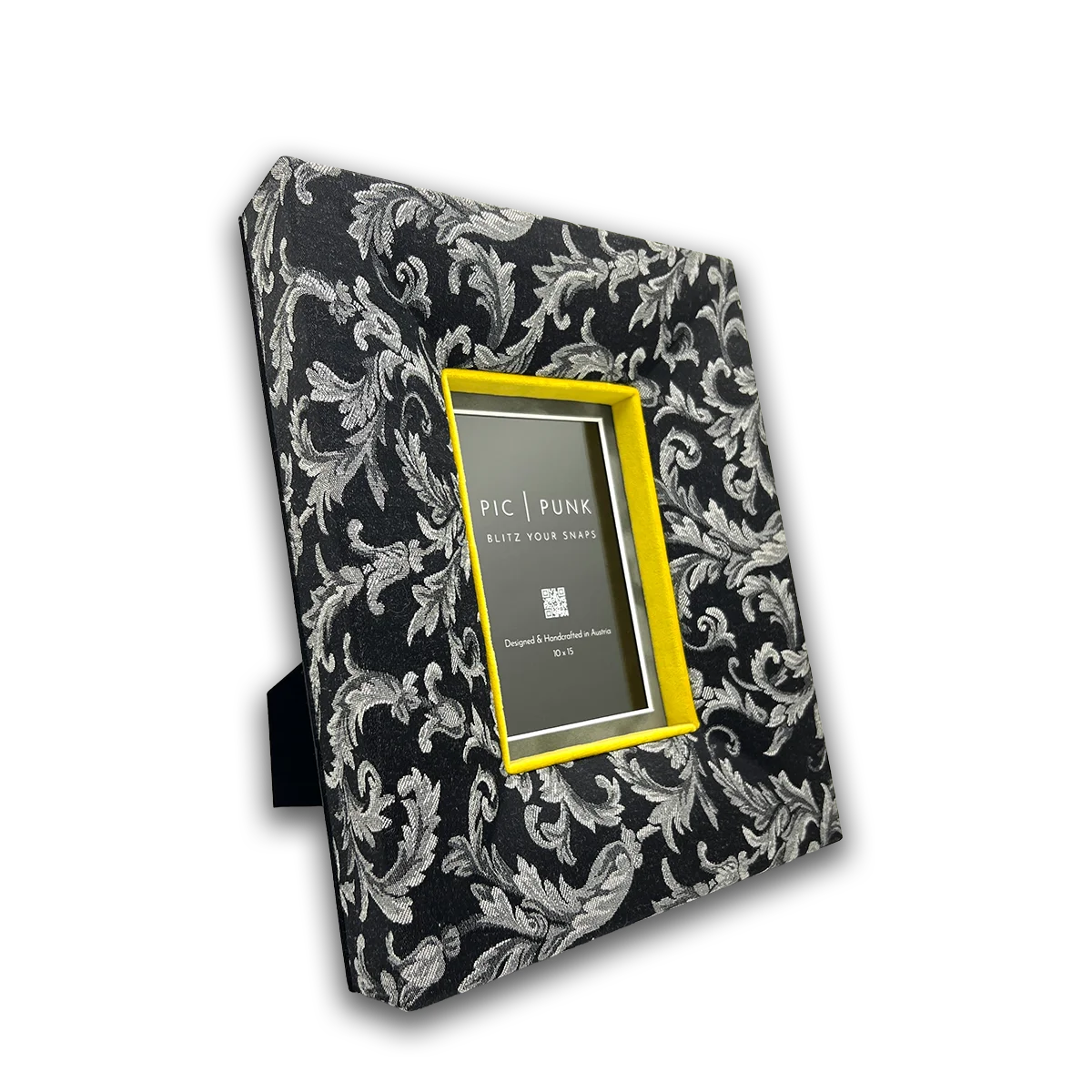 Schräg aufgenommener Stoff-Bilderrahmen in Schwarz mit feinem, silbernem Ornamentmuster und leuchtend gelbem Samt-Innenrand. Der handgefertigte Designrahmen steht frei und zeigt seine plastische Tiefe und textile Oberfläche besonders deutlich. Ideal als moderner Fotorahmen für 10×15 cm Bilder, der klassische Muster mit zeitgemäßem Interior-Design verbindet und als Blickfang auf Sideboard, Regal oder Schreibtisch wirkt.
