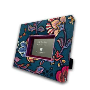 Handgefertigter Stoff-Bilderrahmen von PIC | PUNK® mit kräftigem floralem Muster auf petrolfarbenem Grund, kombiniert mit Akzenten in Beere, Pink, Gelb und Blau. Der tiefe Designrahmen ist mit hochwertigem Textil bezogen und besitzt ein farblich abgestimmtes Samt-Inlay. Schräg dargestellt, zeigt das Bild die markante Rahmenhöhe und die luxuriöse Verarbeitung. Ideal als exklusiver Bilderrahmen für moderne Wohnräume, Galeriewände und individuelle Interior-Konzepte.