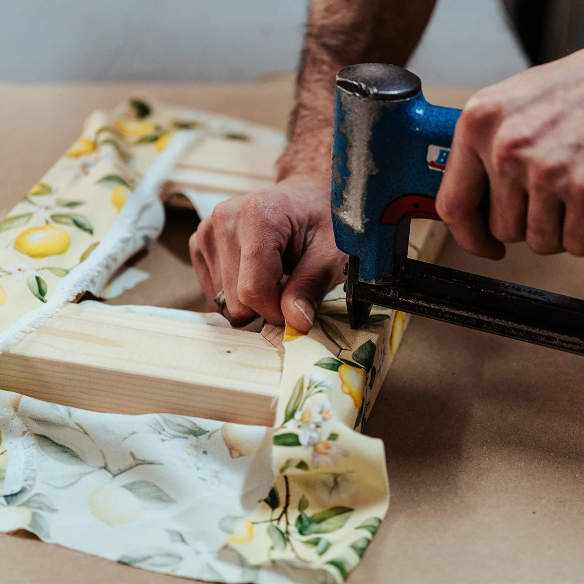 Hände eines Handwerkers spannen einen gemusterten Stoff auf einen Holzrahmen mit Tacker - Herstellung eines stoffbezogenen, handgefertigten Design-Bilderrahmens aus Österreich von PIC | PUNK.