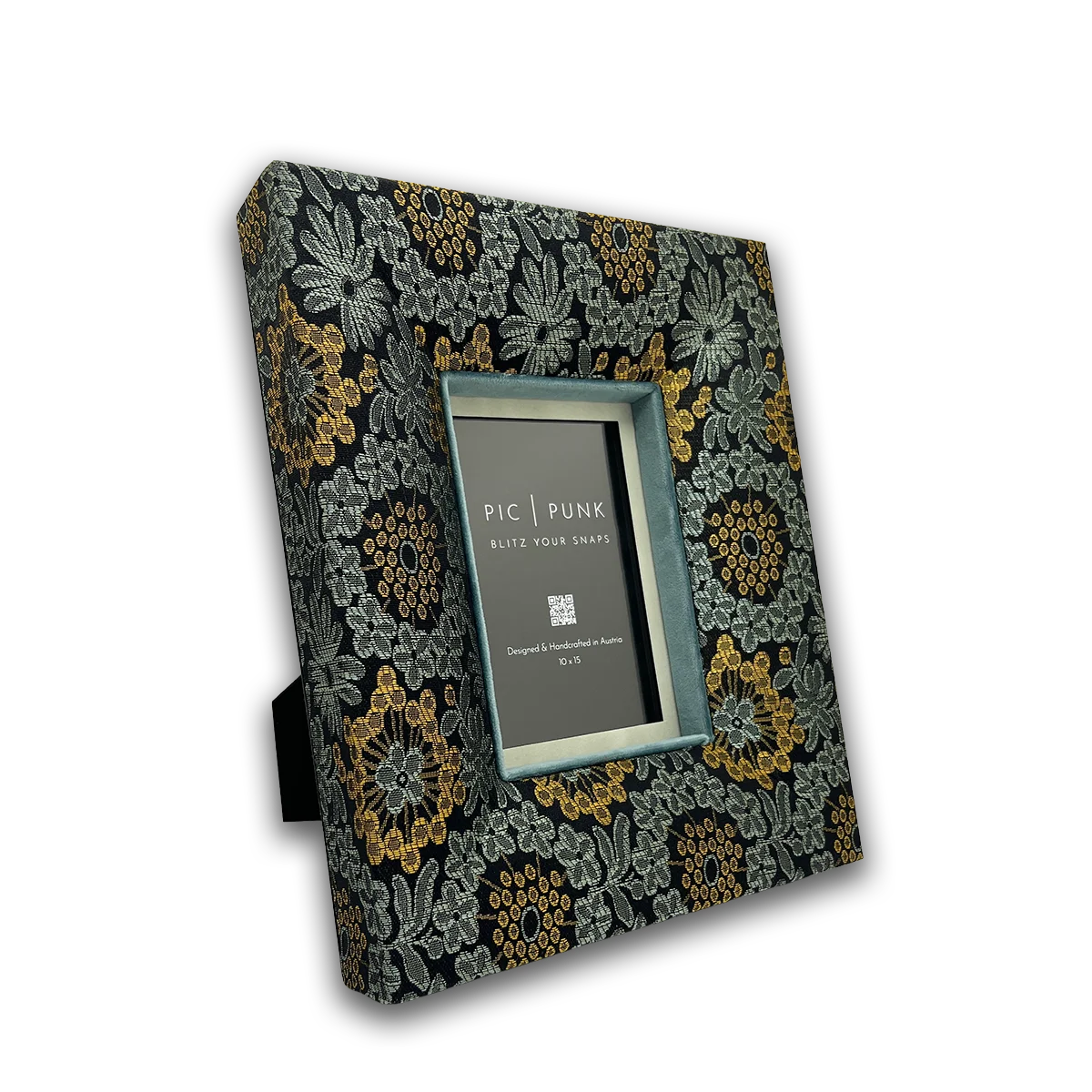 The photo shows the Velvet Underground picture frame from the Soho Style collection. The frame is covered in black fabric, adorned with floral patterns in silver-grey and highlighted with warm mustard-gold accents. The smoky petrol-blue velvet inlay adds depth and elegance. Handcrafted in Austria, this frame blends urban Soho chic with iconic New York spirit.