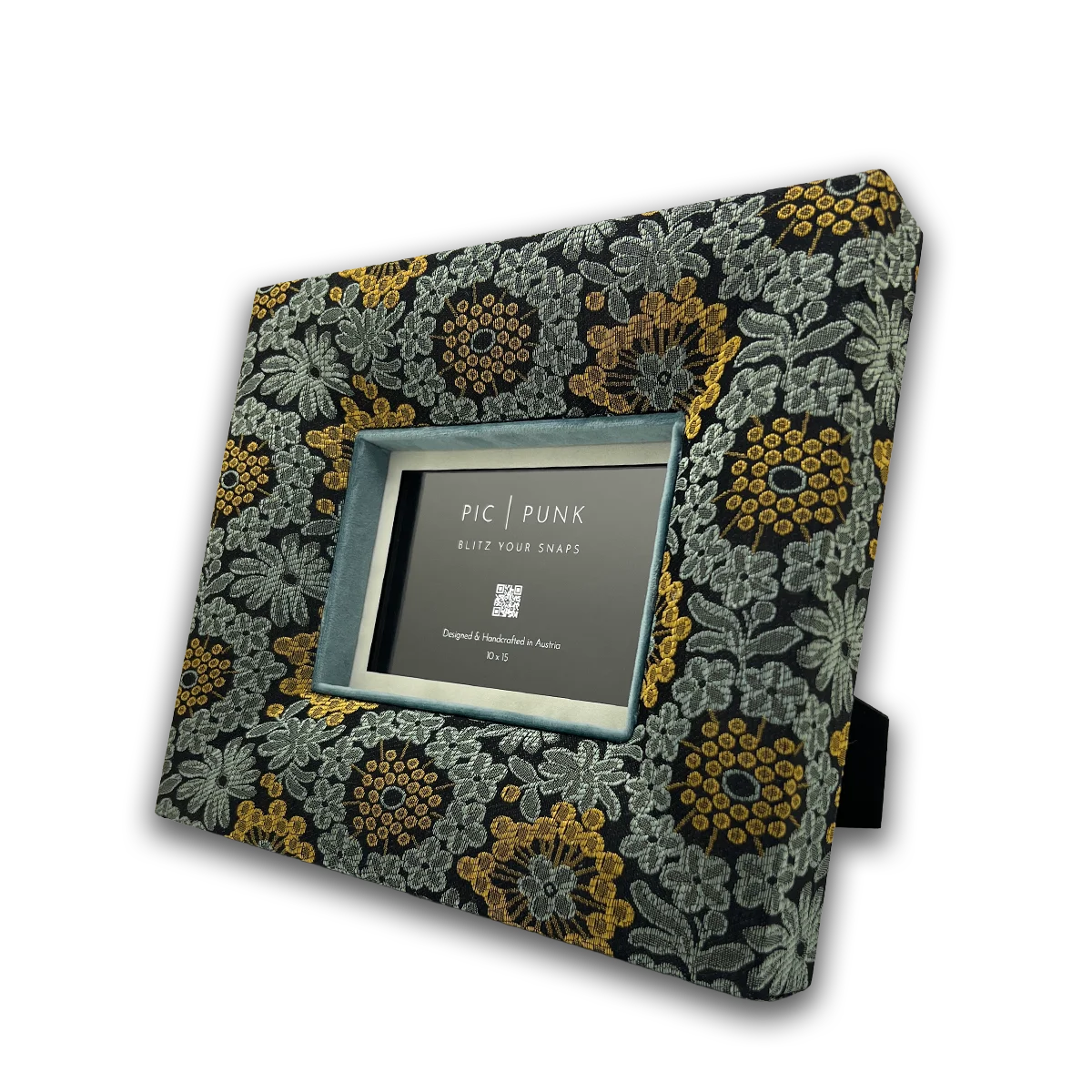 Das Foto zeigt den Bilderrahmen Velvet Underground aus der Kollektion Soho Style. Der Rahmen ist mit schwarzem Stoff bezogen, durchzogen von floralen Mustern in Silbergrau und Akzenten in warmem Senfgold. Das samtige Inlay in Petrolblau verleiht dem Design Tiefe und Eleganz. Handgefertigt in Österreich verbindet der Rahmen urbanen Soho Chic mit einem ikonischen New-York-Feeling.